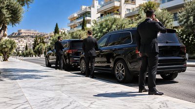 Two plain-clothes close protection officers escort a family from a private residence gate to a waiting armoured sedan on an Athens street.