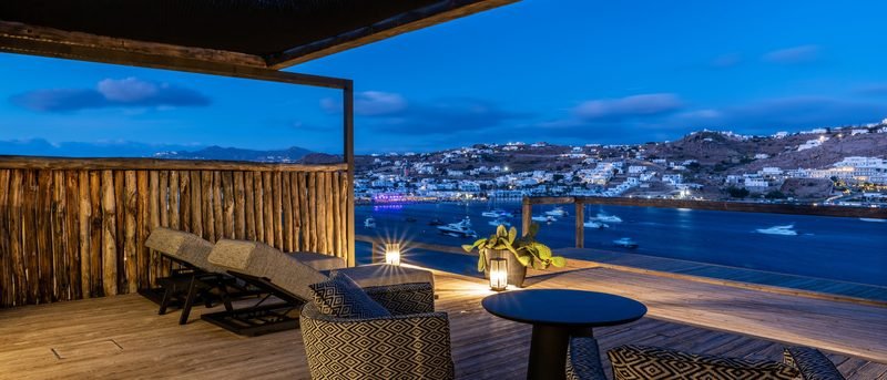 Timber-pergola terrace at blue hour, loungers and lanterns above a boat-filled bay, Mykonos