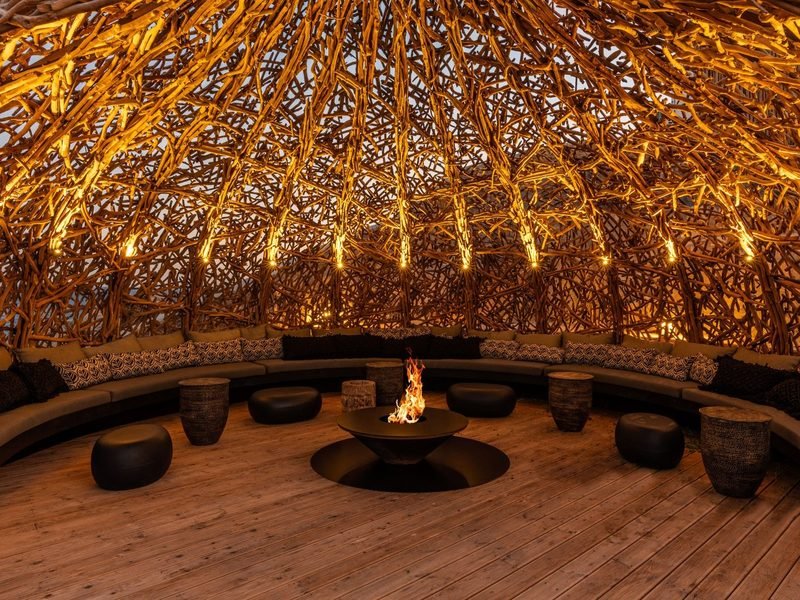 Circular driftwood pavilion with curved banquette seating, woven stools and a burning central fire bowl