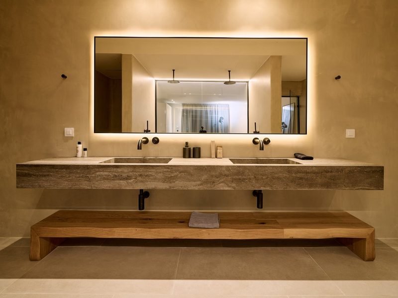 Double stone vanity, backlit mirror and live-edge oak bench against warm plaster walls