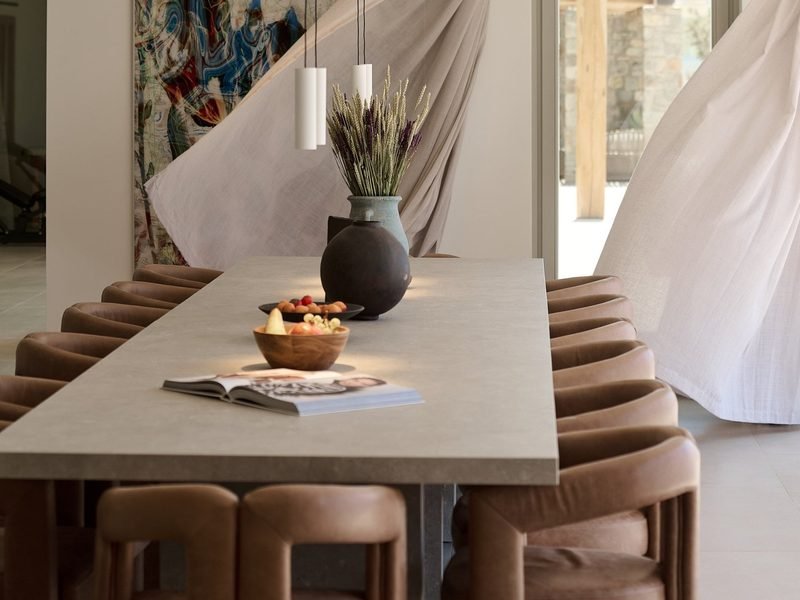 Long dining table laid with fruit bowls and dried stems between tan leather chairs