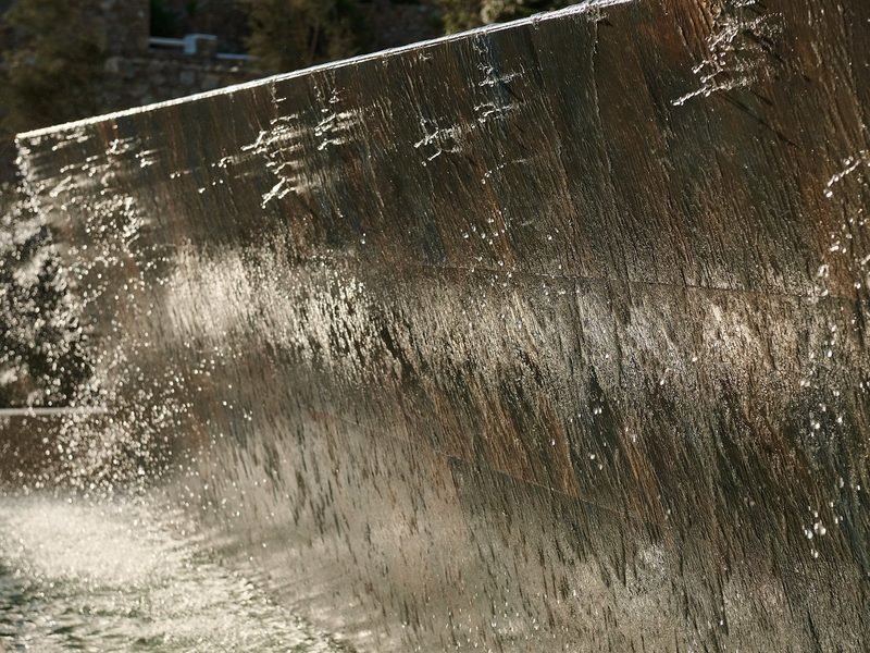 Water sheets down a dark stone wall, sunlight catching the falling spray