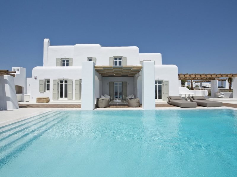 Whitewashed villa with grey shutters seen across the still pool in full daylight