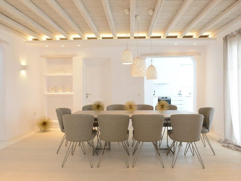 Long dining table ringed with upholstered chairs under woven pendant lamps and beamed ceiling