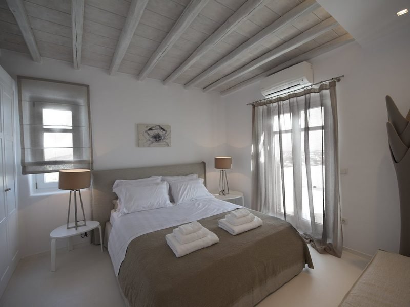 Double bed dressed in white and taupe below a beamed ceiling, curtained window