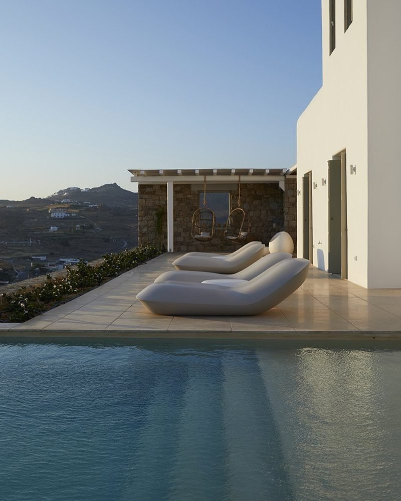Poolside terrace with curved loungers and stone pergola beside the villa's white volume