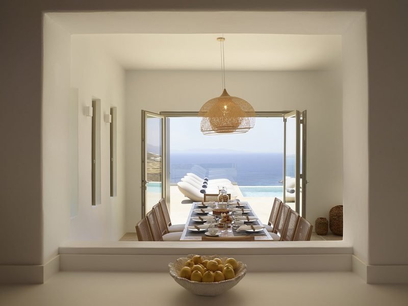 Dining table laid with plates and glasses beneath a woven pendant lamp, sea beyond