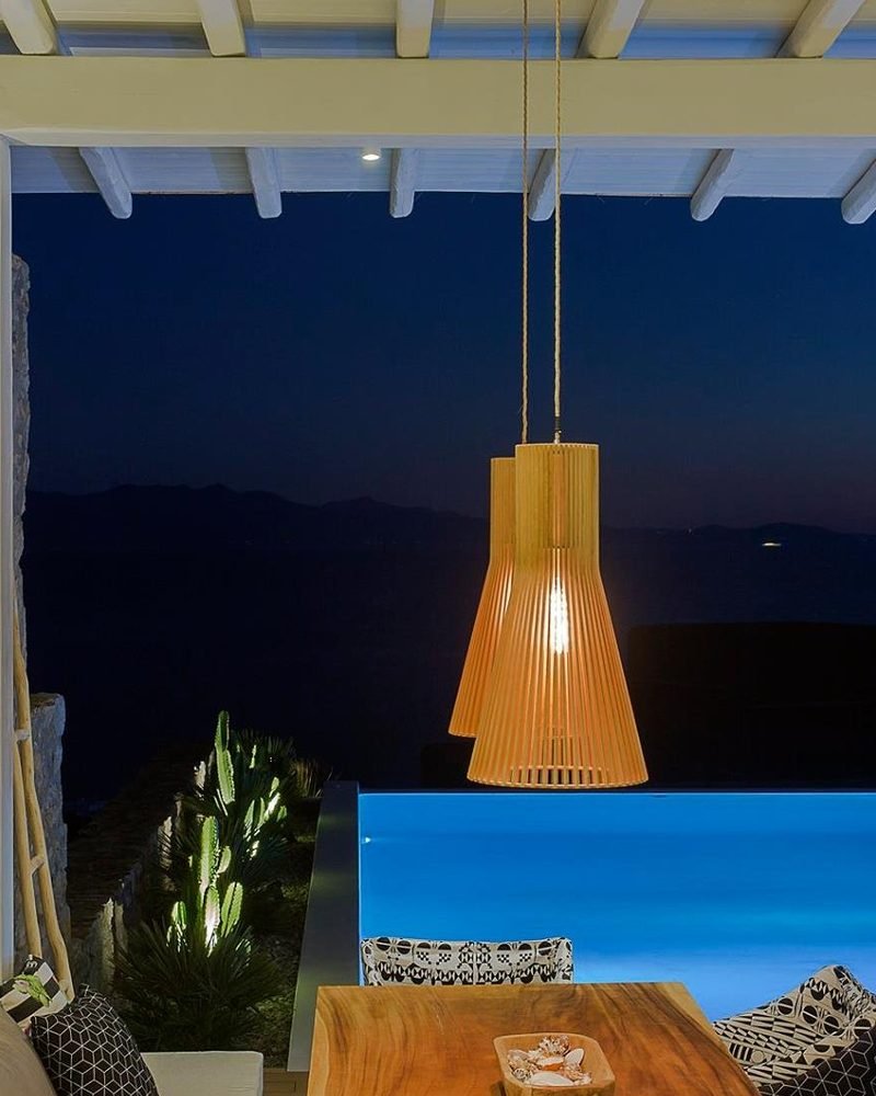 Pergola dining table under a woven pendant lamp beside the lit infinity pool at night