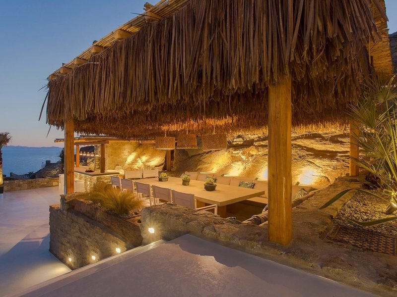 Long outdoor dining table under a thatched reed canopy carved into the rock face