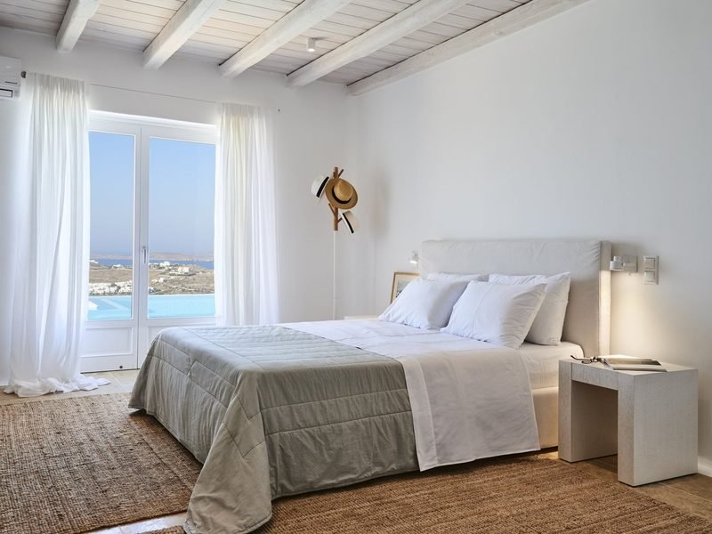 Bedroom in white with beamed ceiling, balcony doors opening toward pool and sea