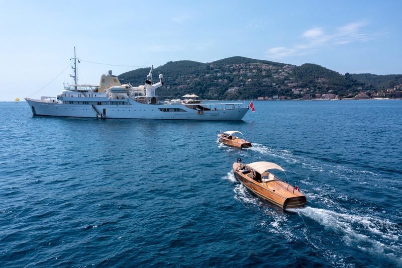 White motor yacht at anchor off a wooded headland, two mahogany tenders running past