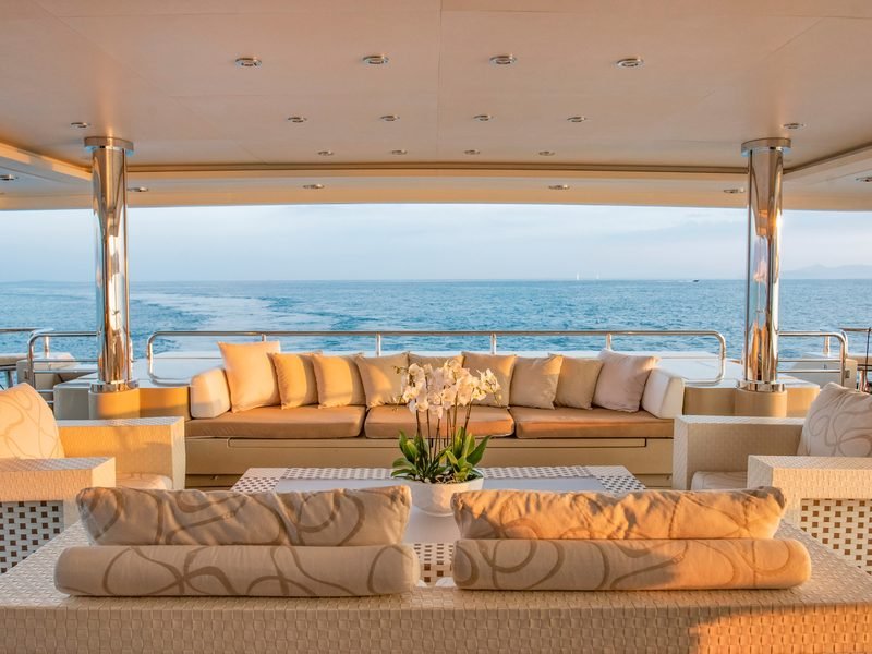 Shaded aft-deck lounge at golden hour, long banquette facing the wake astern
