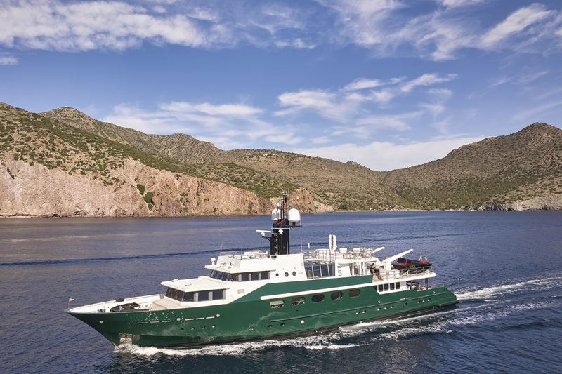 Dark green motor yacht underway along an arid Greek coastline, port profile