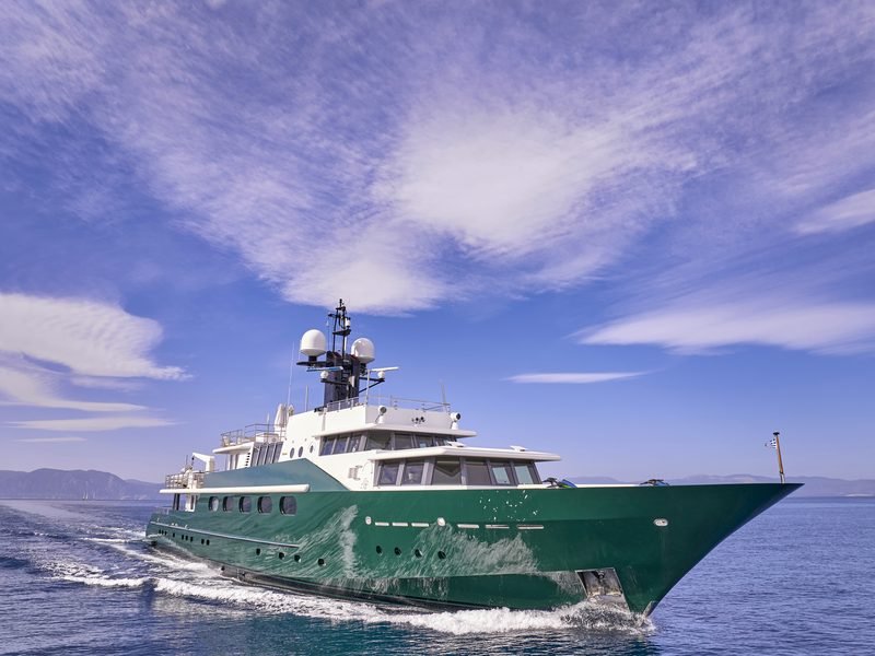 Bow three-quarter view underway, dark green hull and Greek ensign on calm sea