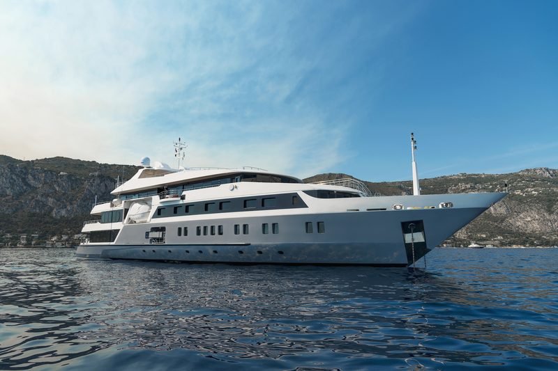 White five-deck motor yacht at anchor below a rocky Riviera headland