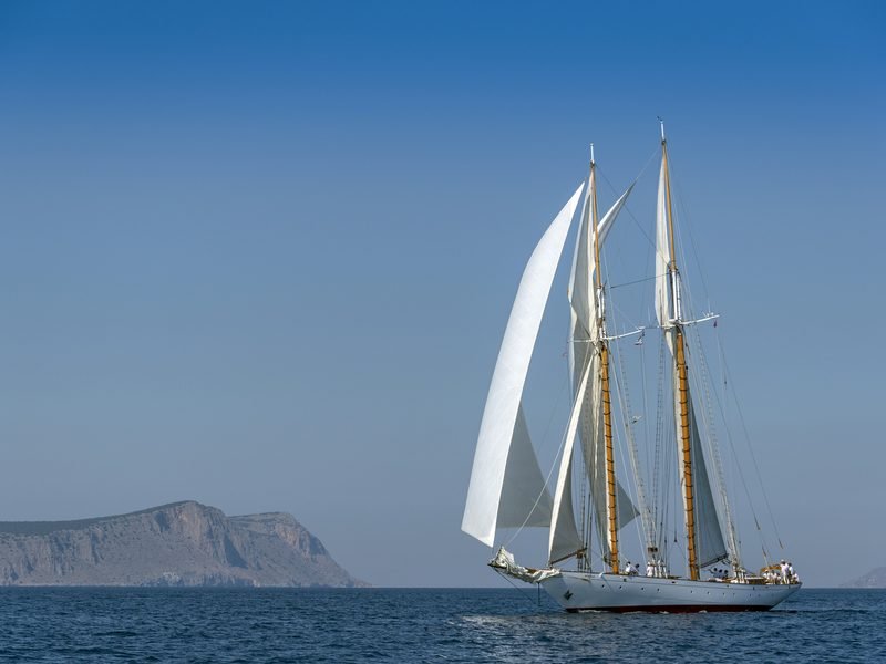 Puritan under sail on calm water, a rocky Greek headland beyond her bow