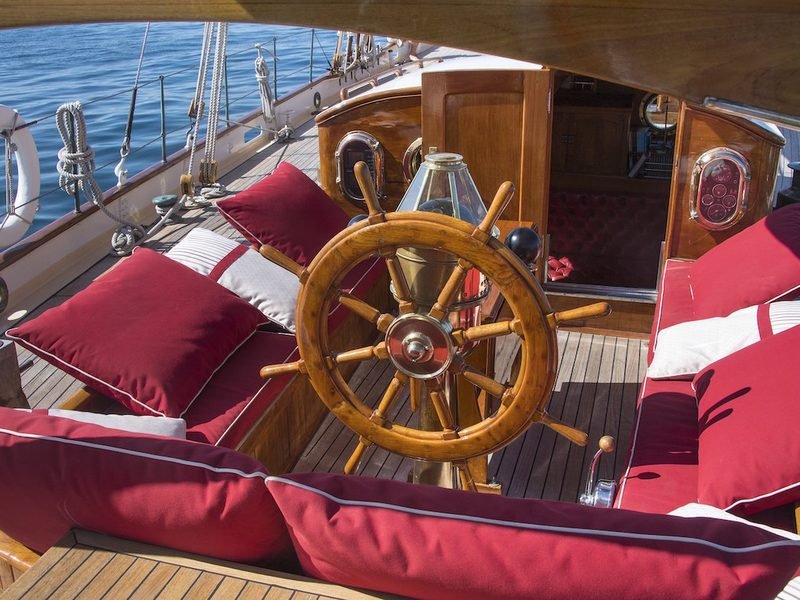 Gaff-era wheel and brass binnacle in the after cockpit, oxblood cushions and varnished deckhouse