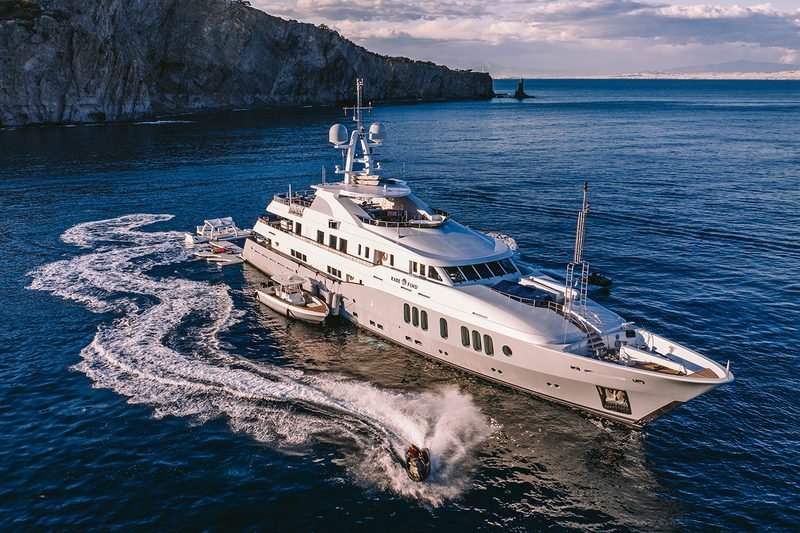 White motor yacht at anchor below a rocky headland, jet ski carving past alongside