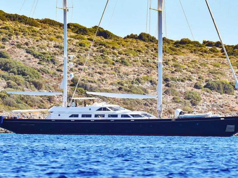 Full port profile at anchor below a scrubland headland, ketch rig furled